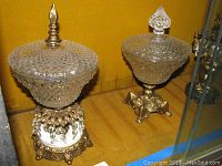 Two cut glass candy dishes with brass bases and finials on lids displayed on yellow cloth.