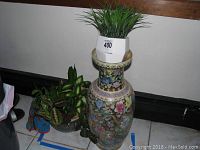Tall oriental ceramic vase with intricate floral and bird designs, with white hexagonal pot containing faux grass plant placed on top.