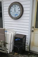 Outdoor wall with 25-inch diameter clock and thermometer mounted, with metal ash can below it and a metal plant stand in front.