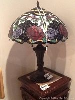 Full view of the Tiffany style stained glass lamp on a wooden table showing the floral and grape motif on the shade and the dark metal base.