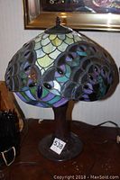Photo showing full view of Tiffany style lamp with stained glass shade and dark brown textured base, placed on a wooden surface with a number tag '538'.