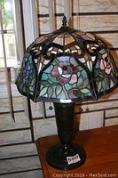 Full view of the Tiffany style lamp showing the stained glass rose design shade and dark metal base on wooden table.