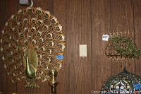 Overall wall view of decorative metal peacock with fan-shaped tail feathers mounted on wood paneled wall next to another metal wall piece.
