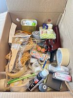 Cardboard box overview showing tapes, paint brushes, twine, paint bottle, knife, notebook, tape dispensers, pen set, paper clip case