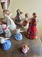 Group shot showing variety of angel figurines including tall red robed angel and multiple small blue and pink kneeling angels
