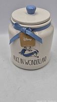 Front view of cream Rae Dunn Disney Alice in Wonderland ceramic canister with blue ribbon and lid knob