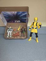Open blister tray showing Buzz Lightyear figure, female ranger figure, yellow rifle, orange blaster, crab creature; yellow-masked figure standing beside tray