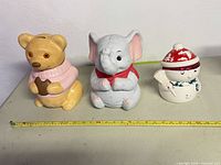 Front view of three ceramic cookie jars: teddy bear, elephant, snowman with measuring tape for scale
