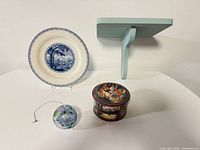 All four items arranged: plate on stand, glass ornament, illustrated tin with lid, painted wood shelf