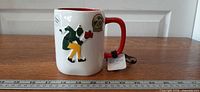Full mug showing raised elf design, red handle, ruler for scale