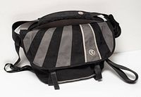 Front view of closed messenger bag showing grey diagonal panels, reflective stripe, logo badge, shoulder strap and pad