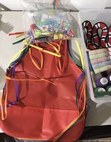 Red craft apron with ribbon ties and bag of beads, pom-poms and pipe cleaners