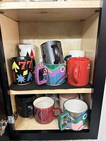 Cupboard shelf showing multiple ceramic mugs with various designs