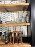 Shelved assortment of clear bowls, ribbed tumblers, footed mug, packaged pink-stem wine glasses, smoky grey stemless glasses, black bowls