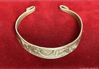 Front view of gold-tone cuff bracelet on red background showing engraved scroll design