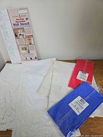 Packaged wall stencil, three folded embroidered linens, two sealed plastic tablecovers red and blue