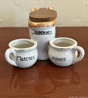 Front view of ceramic novelty ashtray showing Cigarettes jar with lid and side cups labeled Matches and Ashes