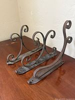 Three wrought iron scroll shelf brackets aligned on wooden surface.
