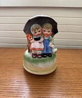 Front view of ceramic music box displaying boy, girl, goose, umbrella and grassy base