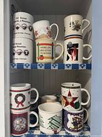 Top two shelves of narrow cabinet showing nine ceramic mugs with Girl Scout, World's Greatest Teacher and Christmas motifs