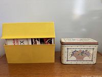 Yellow plastic recipe card box next to cream metal tin