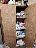 Tall light oak cabinet open showing shelves with jars, books, craft supplies; ceramic duck, vases and travel mug on top
