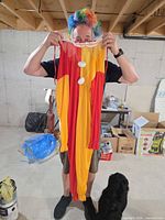 Person holding red/yellow clown jumpsuit front view with white pom-poms and wearing rainbow wig