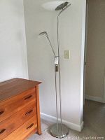 Full view of tall silver-tone metal floor lamp with two lights including torchiere top light and adjustable smaller reading light; lamp placed beside a wooden dresser.