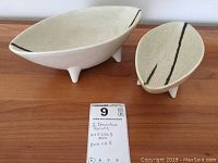 Pair of two decorative white oval stoneware bowls with three small feet and black line accents, shown on wood surface with measurement card.