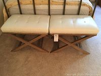 Two ivory faux leather benches with button tufted tops and wooden X-shaped bases placed on carpet in front of a bed.