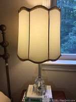 Full lamp showing tall scalloped silk shade with gold trim and lit base on wooden table near window