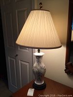 Full view of the lamp lit showing pleated fabric shade, porcelain body with floral design and black base.