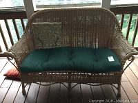 Front view of the wicker settee with green cushion and decorative throw pillow on a wooden deck.