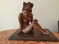 Front view of the clay sculpture seated young nude girl on wooden base, showing detailed posture and bow in hair.