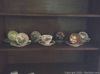 Photo of seven teacups on top of matching saucers with various floral and fruit designs displayed on a wooden shelf.
