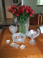 Eight cut glass pieces arranged on a tabletop including a heart shaped bowl, a large footed bowl, a smaller footed bowl, two small salt and pepper cellars, and a rectangular tray, with a vase holding red flowers behind them.