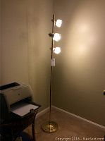 Full view of the 63 inch brass floor lamp standing in a corner beside a desk and printer, illuminated and showing its three light sockets with mesh shades.