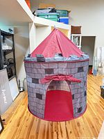 Exterior view of red and gray castle play tent showing conical roof, brick pattern walls and doorway flap
