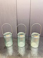 Three ribbed light-green glass lantern candle holders with metal handles displayed upright