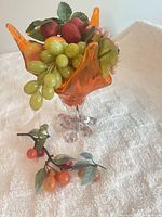 Front view of orange art glass vase filled with assorted artificial fruit, extra loose fruit stems beside