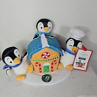 Front view showing three penguins and gingerbread house plush with attached tag