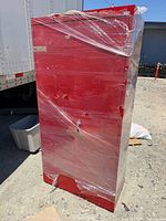 Front view of red metal cabinet wrapped in stretch film showing handle, lock, holes, lower dent