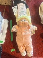 Full view of blonde Cabbage Patch Kids baby doll with overalls, papers and tag visible