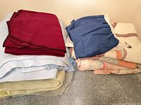 Stacked burgundy, blue, green blankets and navy bed skirt next to folded shell bedspread