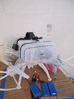 Front and top view of white quadcopter drone with camera and VR headset on top, batteries and charger in front