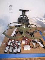 Overall lot showing Kolibri olive green drone with propeller guards, FPV remote control behind it, extra white and black blades around, batteries and charging hubs on tabletop.