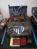 Photo showing multiple purses and bags including a large black tote, small brown purse, medium black zippered bag, and a large black bag with a heart design, along with several wallets in front.
