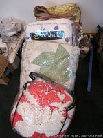 Pile of blankets and quilts including a white quilt with green patchwork and a red and white crochet blanket in plastic bag