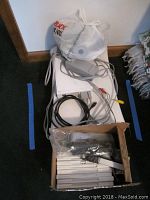 Nintendo Wii console laying flat with power cable, AV cables and sensor bar, controller accessories inside a box, and a white plastic bag presumed to contain more accessories.