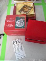 Photo showing three Christmas ornaments and their original boxes on a table, including a red velvet box, a gold box with a clear lid, and a green Mikasa box.